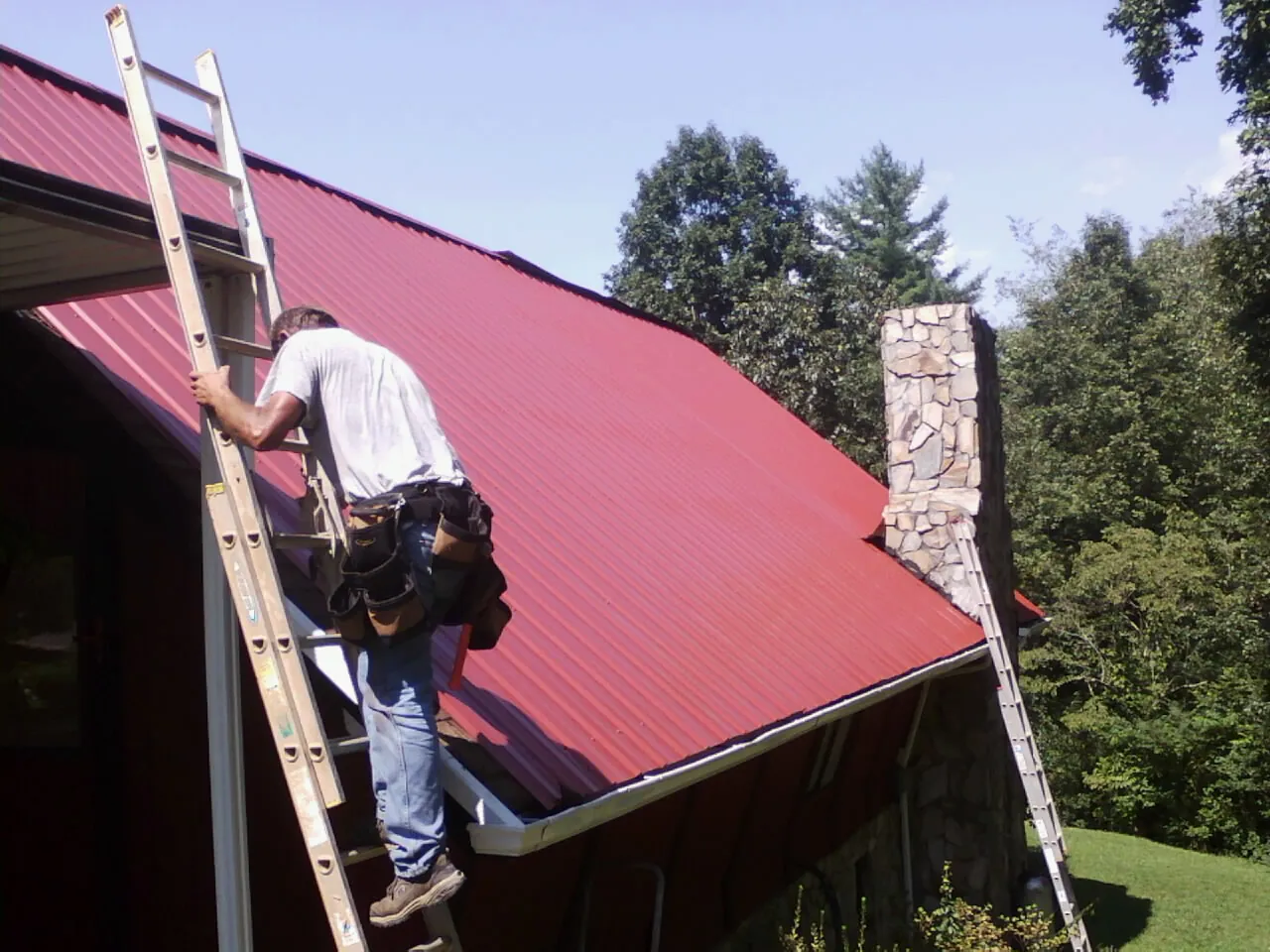 Roofer on a ladder beside a red metal roof and stone chimney — Soffit & Fascia Repair by Roofing Brothers in Smithsburg